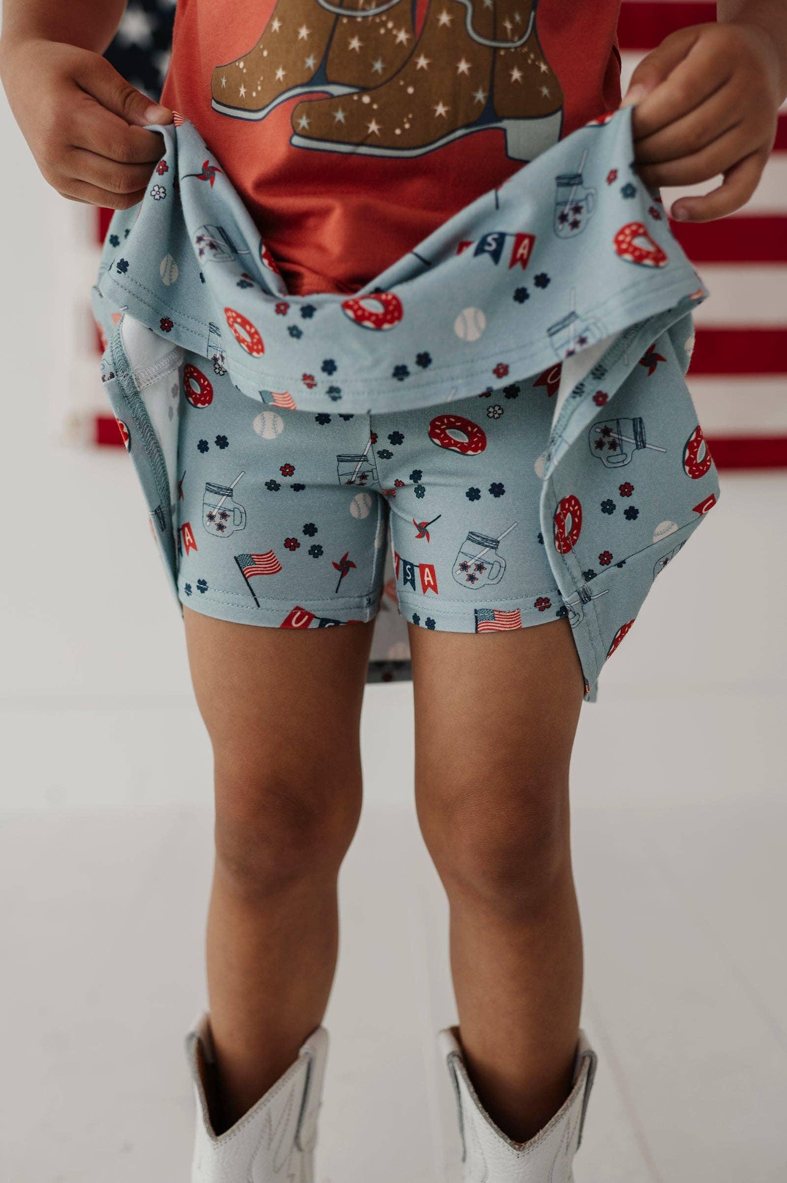 Child wearing patriotic print shorts with donuts, flags, and mason jars, paired with white boots