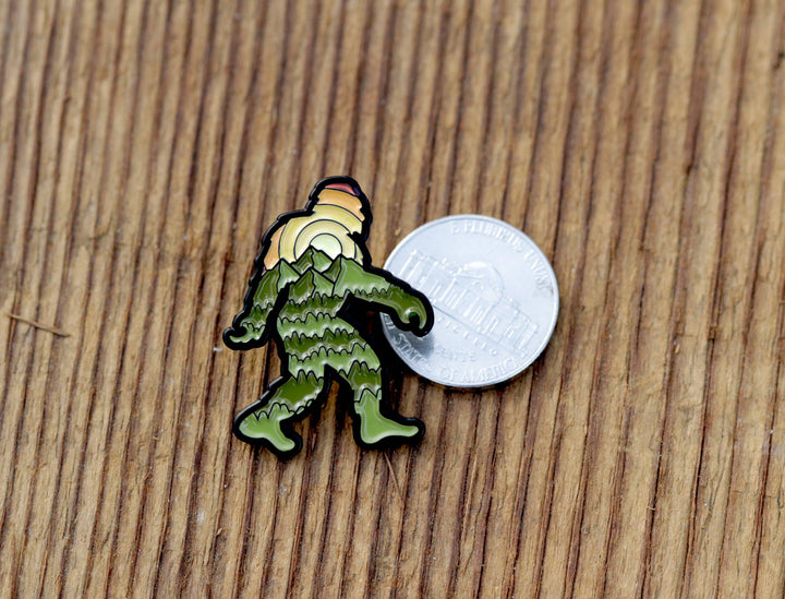 Green mountain-themed enamel pin with hiker design, shown next to a quarter for scale on wood.
