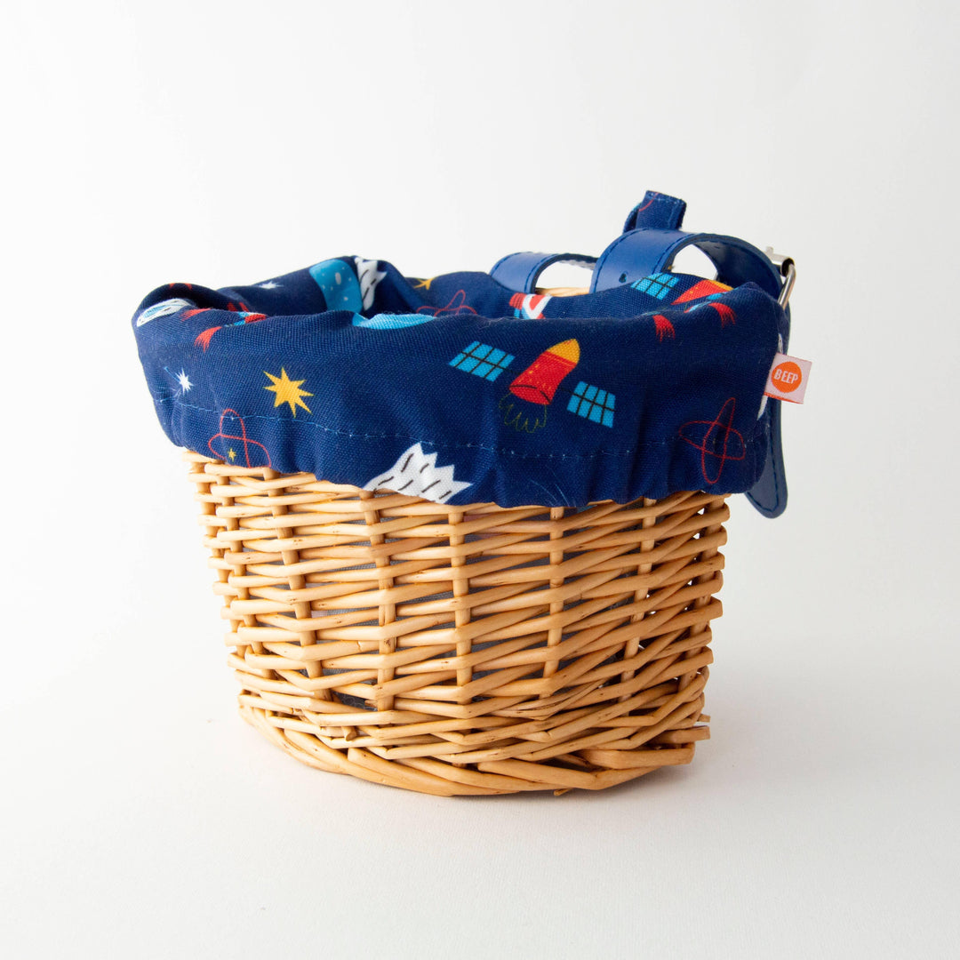 Wicker bike basket with space-themed navy liner, rockets, stars, and BEEP tag for outdoor adventures.