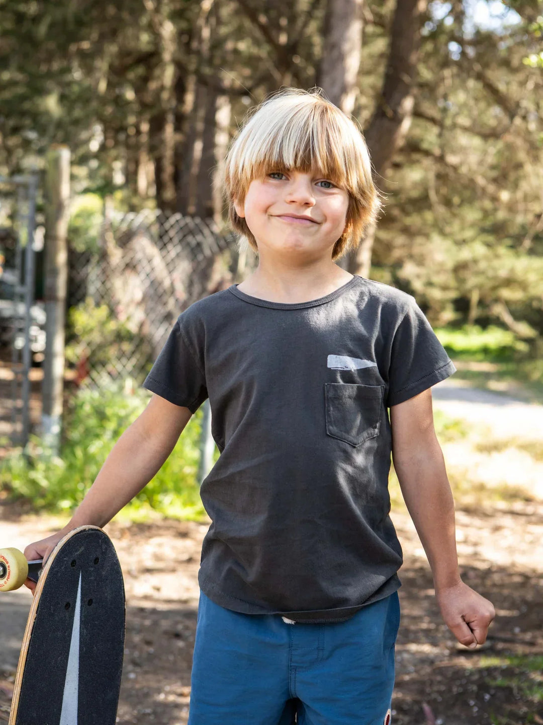 Kid holding skateboard outdoors in PNW forest, wearing dark tee and blue shorts for coastal adventure gear