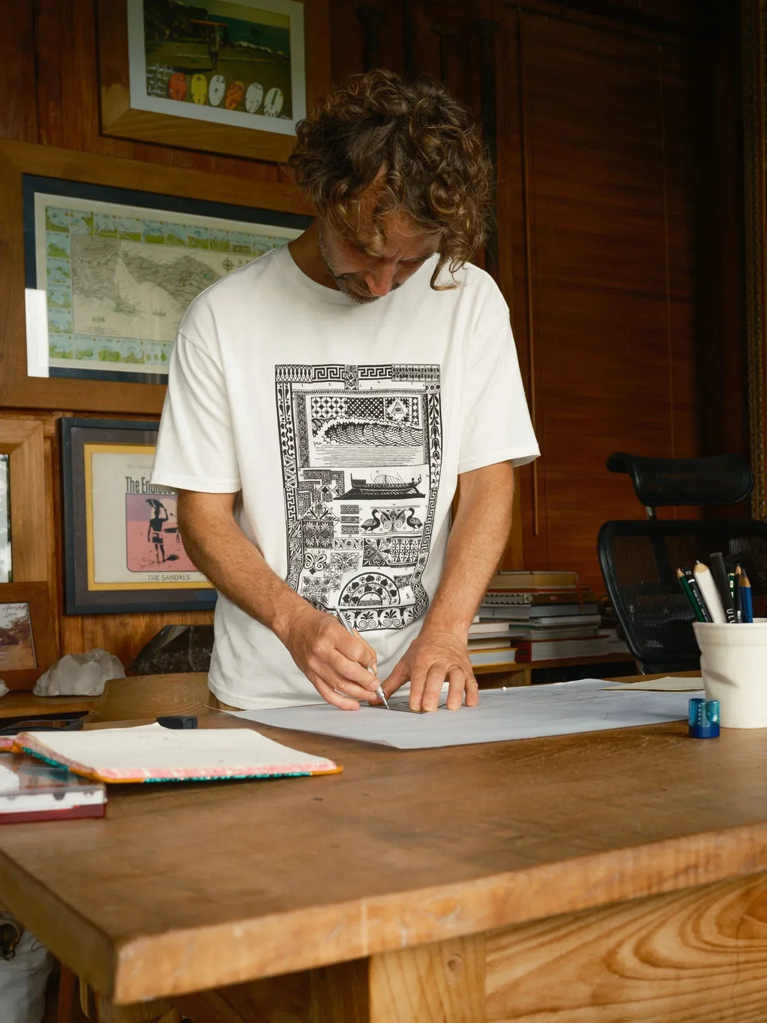Man in white graphic tee sketching at wooden desk, coastal art on walls, PNW outdoor lifestyle