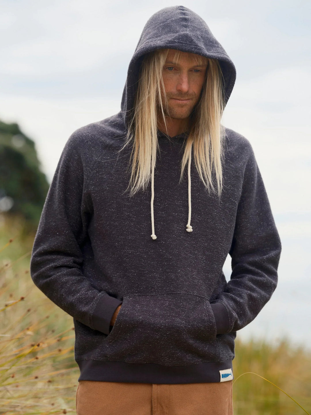 Man in navy speckled hoodie with drawstring hood, hands in pocket, coastal dunes background
